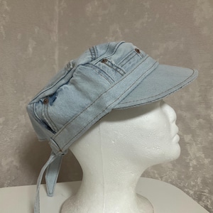 May include: A light blue denim military-style cap featuring a structured brim, visible copper-toned metal rivets, and stitched paneling. The hat includes a fabric strap detail at the back and is displayed on a white mannequin head.