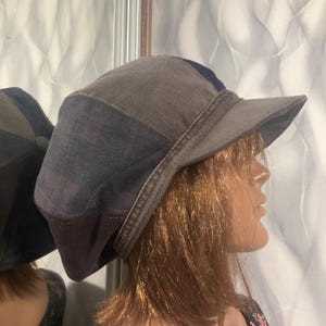 Puede incluir: Un sombrero de mezclilla patchwork en tonos grises, azules y morados. El sombrero presenta una visera corta y una corona redondeada. El sombrero se exhibe en una cabeza de maniquí.