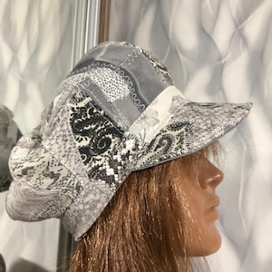 Puede incluir: Una gorra estilo boina de patchwork con una variedad de telas en gris, negro y blanco. El gorro muestra una mezcla de patrones que incluyen cachemir, piel de serpiente y estampados florales, unidos con costuras en zigzag visibles.
