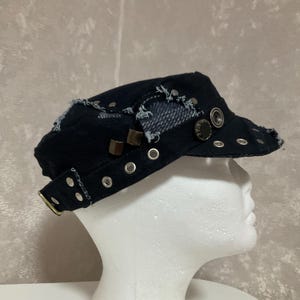 Könnte beinhalten: Eine schwarze Military-Kappe aus Denim mit ausgefransten Kanten, silberfarbenen Metallösen und Zierknöpfen. Die Kappe verfügt über einen seitlichen Riemen mit Metallschnalle und abgenutzten Stoffeinsätzen, präsentiert auf einem weißen Mannequinkopf.
