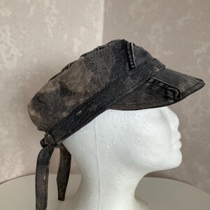 May include: A charcoal grey and black acid-wash denim cap featuring a structured visor and a tie-back closure. The fabric displays a mottled, distressed pattern throughout, with stitched seams along the crown and a wide band encircling the base.