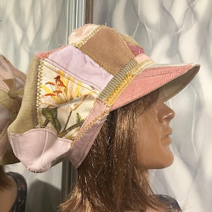 May include: A patchwork newsboy cap constructed from various fabric textures including tan corduroy, light pink denim, and a floral print panel featuring yellow lilies. The panels are joined with visible yellow zigzag stitching and a pink brim.
