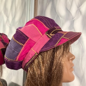 Puede incluir: Una gorra tipo boina de patchwork con paneles alternos de tela morada con un sutil patrón floral y tela rosa brillante sólida. Las secciones están unidas por costuras en zigzag amarillas visibles y la gorra incluye una visera rígida.