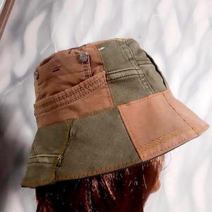 May include: A patchwork bucket hat in shades of brown and olive green. The hat features a mix of fabrics and textures, with visible stitching and metal button details. The brim is a solid brown color.