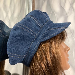 Puede incluir: Gorra de estilo newsboy de mezclilla azul medio. La gorra presenta un diseño estructurado con una visera corta y costuras visibles en un color beige contrastante. Un estilo clásico, adecuado para varios atuendos informales.