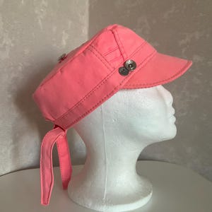 May include: A bright coral pink military-style cap featuring a structured crown, a stiff brim, and two silver-toned metal buttons on the side. The cap includes two fabric ties hanging from the back and visible topstitching throughout the design.