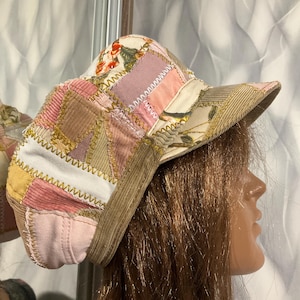 May include: A patchwork newsboy cap with a corduroy brim. The cap features a variety of fabrics in shades of pink, beige, and floral patterns. The hat is constructed with visible stitching and has a rounded crown.