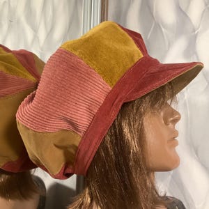 May include: A multi-colored newsboy cap crafted from corduroy fabric. The hat features alternating panels in shades of mustard yellow, dusty pink, and deep red. It includes a structured brim and a rounded crown, showcasing a textured, ribbed surface throughout the design.