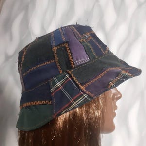 Könnte beinhalten: Ein Patchwork-Eimerhut mit verschiedenen Stoffen in Blau-, Grün- und Lilatönen. Der Hut hat eine Krempe und ist mit sichtbaren Nähten aus kontrastierendem orangefarbenem Faden versehen. Das Design umfasst karierte und unifarbene Stoffflicken.