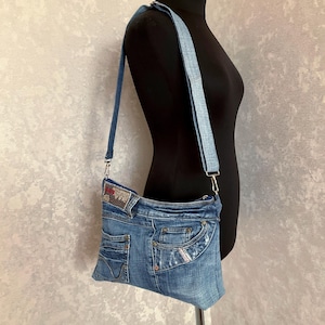 Puede incluir: Un bolso de hombro de mezclilla azul confeccionado con tela de jeans reciclada, con bolsillos visibles, remaches metálicos y una correa de mezclilla a juego. El bolso incluye un pequeño parche rectangular con un diseño estampado en la esquina superior izquierda.