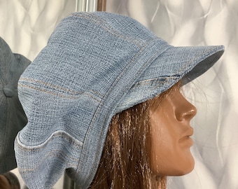 May include: A light blue denim newsboy cap featuring a structured brim and a multi-paneled crown with visible tan stitching. The fabric has a textured weave, and the cap is designed with a rounded, slightly slouchy silhouette that sits on the head.