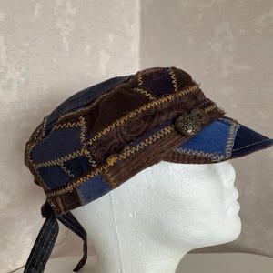Könnte beinhalten: Eine Patchwork-Militärmütze aus verschiedenen blauen und braunen Cord- und Stoffbahnen, die mit sichtbaren gelben Zickzacknähten verbunden sind. Die Mütze verfügt über einen festen Schirm und zwei dekorative Metallknöpfe an der Vorderseite.