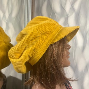 May include: A mustard yellow corduroy newsboy cap featuring a wide, rounded crown with vertical ribbed texture and a stiff, protruding visor. The hat is constructed from soft, thick fabric with a structured band that sits around the head.
