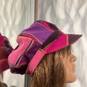 Puede incluir: Una gorra de patchwork confeccionada con varios paneles de tela en tonos morado, magenta y violeta oscuro. El sombrero presenta costuras en zigzag amarillas prominentes a lo largo de las uniones, una corona estructurada y una visera rígida.