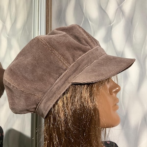 May include: A brown corduroy newsboy cap with a short brim. The hat has a rounded crown and a band around the base. The fabric has a ribbed texture and is a muted brown color.
