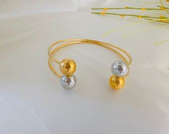 Könnte beinhalten: Ein goldenes Armreif-Armband mit zwei runden Perlen, eine goldene und eine silberne.