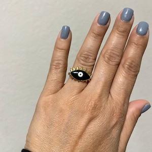 Puede incluir: Un anillo de oro con un diseño de ojo mal de ojo negro. El ojo tiene una pupila blanca y está rodeado de púas doradas.