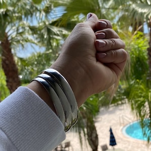 May include: Three silver bangle bracelets stacked on a wrist. The bracelets are smooth and shiny.