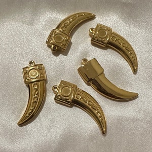 May include: Five gold-colored metal pendants shaped like a curved tusk with intricate designs. Each pendant has a loop at the top for attaching to a chain or cord.
