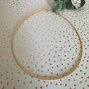 Könnte beinhalten: Eine zarte, goldfarbene Halskette mit einem dezenten, strukturierten Design. Die Halskette ist kreisförmig und scheint aus einem dünnen, flexiblen Material zu bestehen. Der Hintergrund ist hellrosa mit einem Herzmuster und kleinen Zweigen.