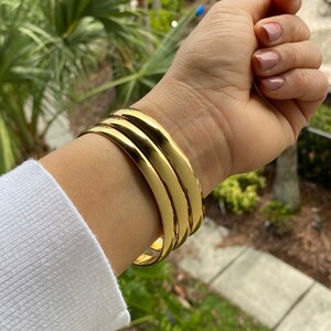 May include: A stack of four gold-colored bangles. The bracelets are smooth and shiny, reflecting light. The jewelry is worn on a wrist, with a white long-sleeved shirt visible in the image.