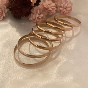 Puede incluir: Un conjunto de seis brazaletes finos de metal en tono oro rosa pulido, dispuestos en una línea superpuesta y en cascada. Las pulseras presentan una superficie lisa y redondeada con un acabado reflectante, creando una forma circular.