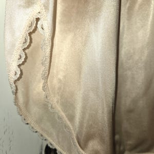 May include: Close-up of a vintage beige slip with delicate lace trim along the edges. The fabric has a smooth, silky texture, and the lace adds a touch of elegance to the garment. The slip appears to be in good condition.