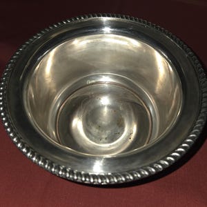 May include: A silver-colored metal bowl with a decorative rope-like trim around the rim. The bowl has a wide opening and a smooth, reflective interior. The bowl is likely used for serving or display.