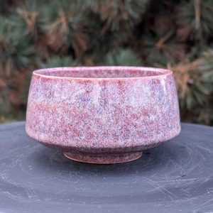 May include: A speckled ceramic bowl with a wide, rounded shape. The bowl has a reddish-purple color with a textured surface. It sits on a small, circular base, and is likely used for serving or display.