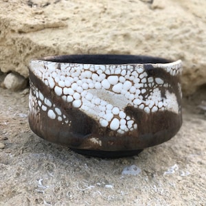 May include: A handmade ceramic bowl with a unique design. The bowl features a dark brown base and rim, with a white and brown speckled pattern on the exterior. The interior is dark brown.