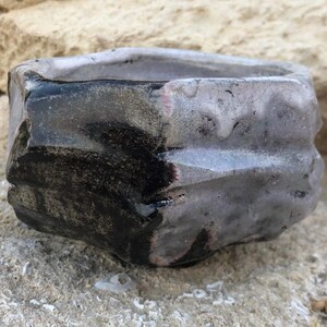 May include: A small, gray and black ceramic pot with a rough, textured surface. The pot has a wide, rounded opening and a slightly pointed base.