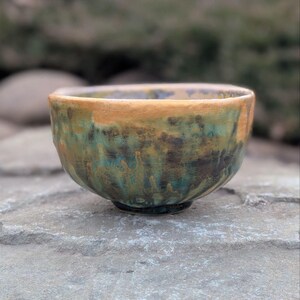 May include: A handcrafted ceramic bowl with a unique glaze. The bowl features a blend of colors, including shades of green, brown, and gold. The exterior has a textured appearance, adding to its artisanal charm. The bowl is suitable for serving or display.
