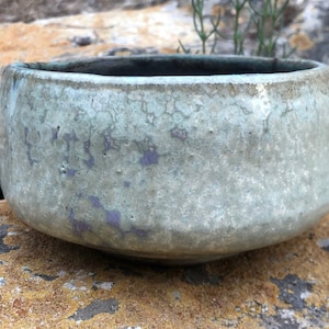 May include: A light green ceramic bowl with a textured surface and a subtle purple glaze. The bowl is sitting on a rock with a rough, textured surface.