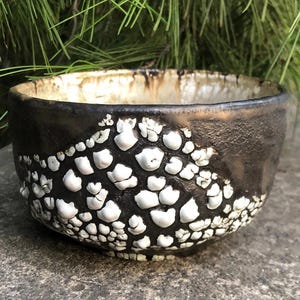 May include: A black ceramic bowl with a textured, white glaze. The bowl has a rounded shape and a wide rim.