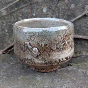 May include: A small, rustic ceramic cup with a light brown glaze and a textured surface. The cup has a slightly irregular shape and a wide rim.
