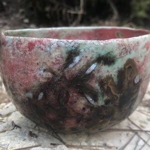May include: A handmade ceramic bowl with a unique, organic design. The bowl features a blend of colors, including red, green, and brown, with white accents. The exterior has a textured appearance, and the bowl is sitting on a stone surface.