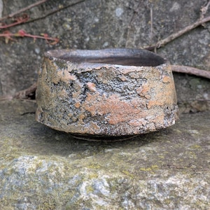 May include: A handcrafted, textured ceramic bowl with a square top and a rounded base. The bowl features a rough, earthy exterior with shades of brown, gray, and hints of orange. The interior is dark, creating a contrast with the textured surface.