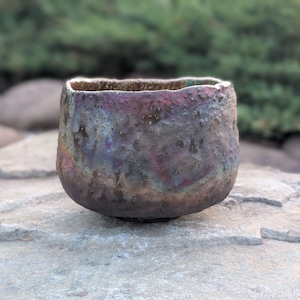 May include: A handcrafted ceramic bowl with a textured surface and a unique iridescent glaze. The bowl features a rounded shape with a slightly irregular rim. The colors include shades of purple, blue, and bronze.