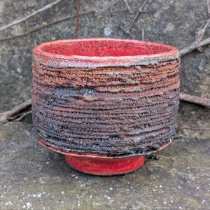 May include: A small, red ceramic pot with a textured, dark brown and black glaze. The pot has a rounded shape and a slightly flared rim.