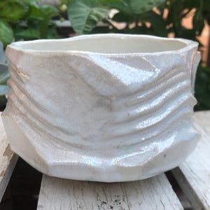 May include: A handcrafted, off-white ceramic planter with a textured, wave-like design. The planter has a wide, open top and a faceted base. The neutral color and unique texture make it a versatile piece for displaying plants or as a decorative item.