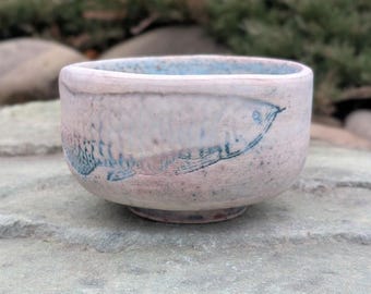 May include: A small, light pink ceramic bowl with a blue fish design. The bowl has a slightly textured surface and a rounded shape.