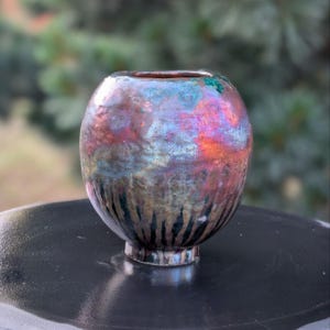 May include: A small, round ceramic vase with a metallic, iridescent finish. The colors shift from red and orange to blue and purple. The base has a dark, textured pattern.
