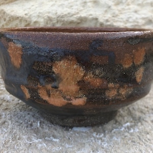 May include: A small, brown ceramic bowl with a dark brown glaze and a speckled, orange-brown design. The bowl is sitting on a rough, gray surface.