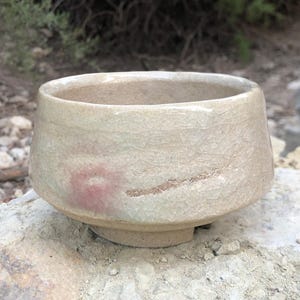 May include: A small, white ceramic bowl with a textured surface and a subtle pink and brown glaze. The bowl has a rounded shape and a slightly flared rim.