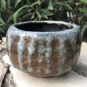 May include: A handmade ceramic bowl with a textured, crackled glaze. The bowl features a blend of brown, gray, and white colors. The interior is dark, and the bowl sits on a stone surface, showcasing its unique, organic form.