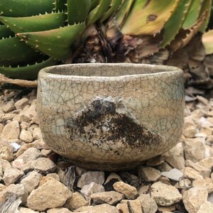 May include: A small, white ceramic bowl with a crackled glaze and a dark brown, textured design. The bowl is sitting on a bed of small, gray rocks.