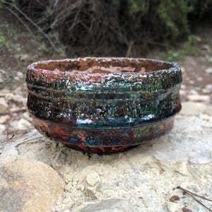 May include: A small, round, ceramic bowl with a dark green, blue, and brown glaze. The bowl has a rough, textured surface and a slightly uneven rim.
