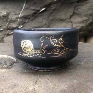 May include: A small black ceramic bowl with a gold snail design. The snail is crawling on a branch with leaves.