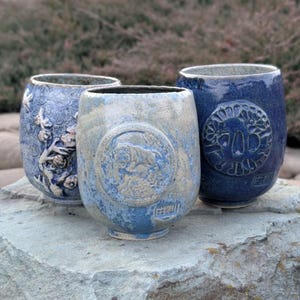 Könnte beinhalten: Drei Keramikbecher mit blau-weißer Glasur. Die Becher haben unterschiedliche Designs, darunter ein Blumenmuster, einen Tiger und eine stilisierte Blume.