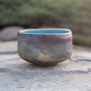 May include: A handmade ceramic bowl with a square shape and a light blue interior. The exterior features a blend of colors, including purple, gold, and gray. The bowl is sitting on a stone surface.